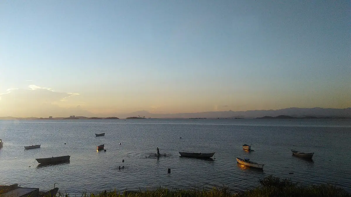 Baía de Guanabara ao entardecer, vista de São Gonçalo, com barcos de pesca na água calma e a serra ao fundo
