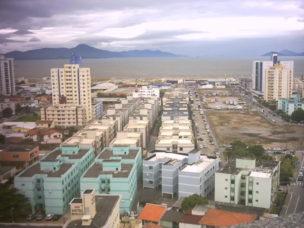 Vista da beira-mar de São José com prédios, a baía sob céu nublado e os morros da ilha de Santa Catarina ao fundo