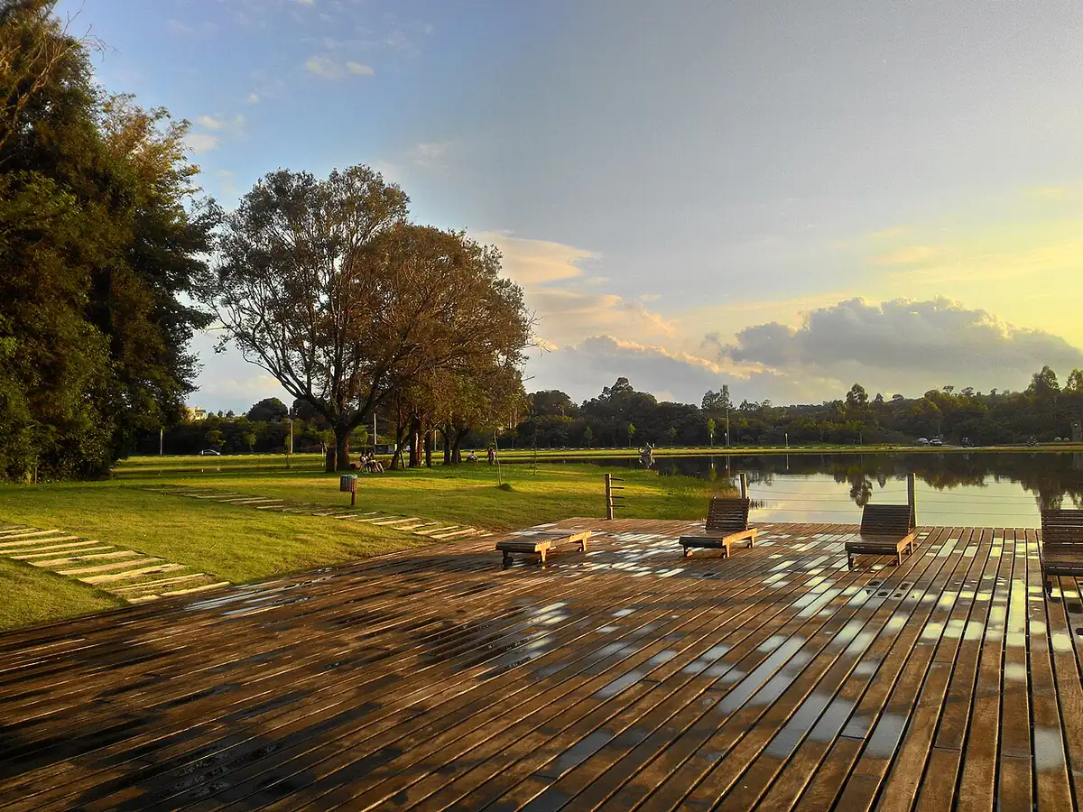 Deque de madeira molhado e gramado às margens do lago do Parque do Povo, em Toledo, com céu de fim de tarde
