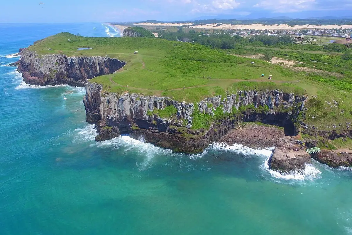 Vista aérea do Morro das Furnas, em Torres, com falésias de rocha caindo sobre o mar verde e as dunas ao fundo