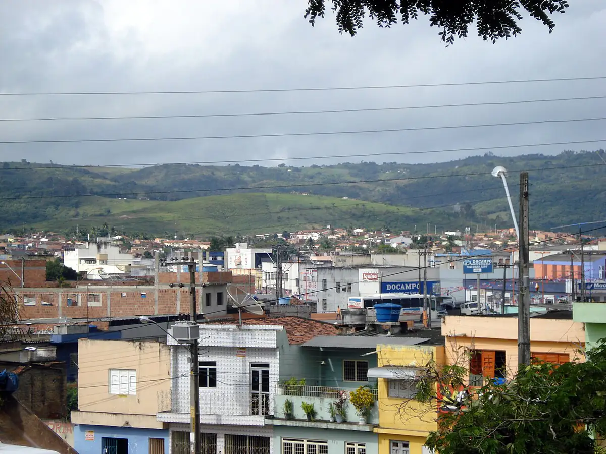 Vista de Vitória de Santo Antão, com casas e prédios do centro e morros verdes ao fundo sob céu nublado